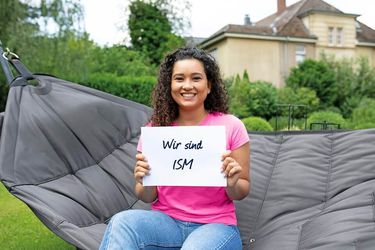Studentin der ISM Fernstudium auf einer Hängematte mit einem Schild, Text: Wir sind ISM